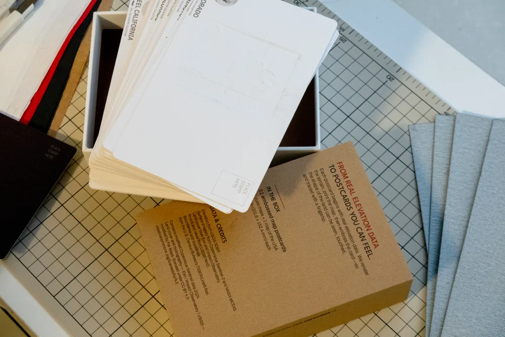 The open box with a stack of proof postcards showing their printed backs, beside the kraft insert listing the box contents and data credits