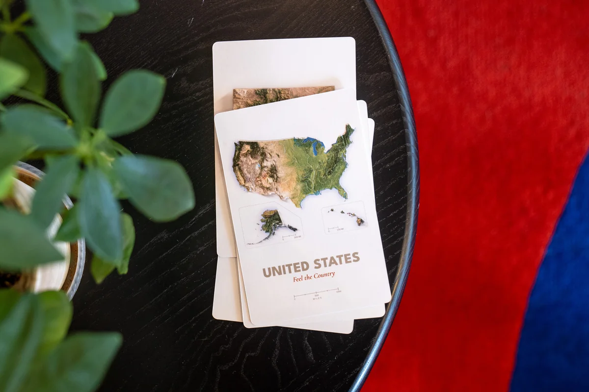 United States relief postcard production proof stacked with other cards on a black table beside a plant