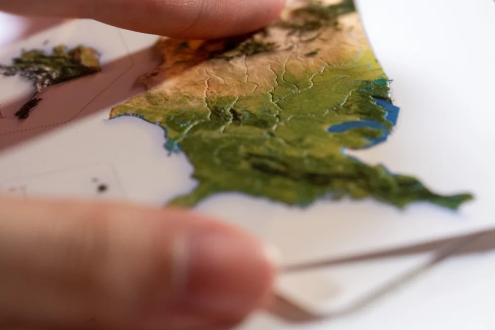 A fingertip tracing the raised western mountain ranges on a United States postcard production proof