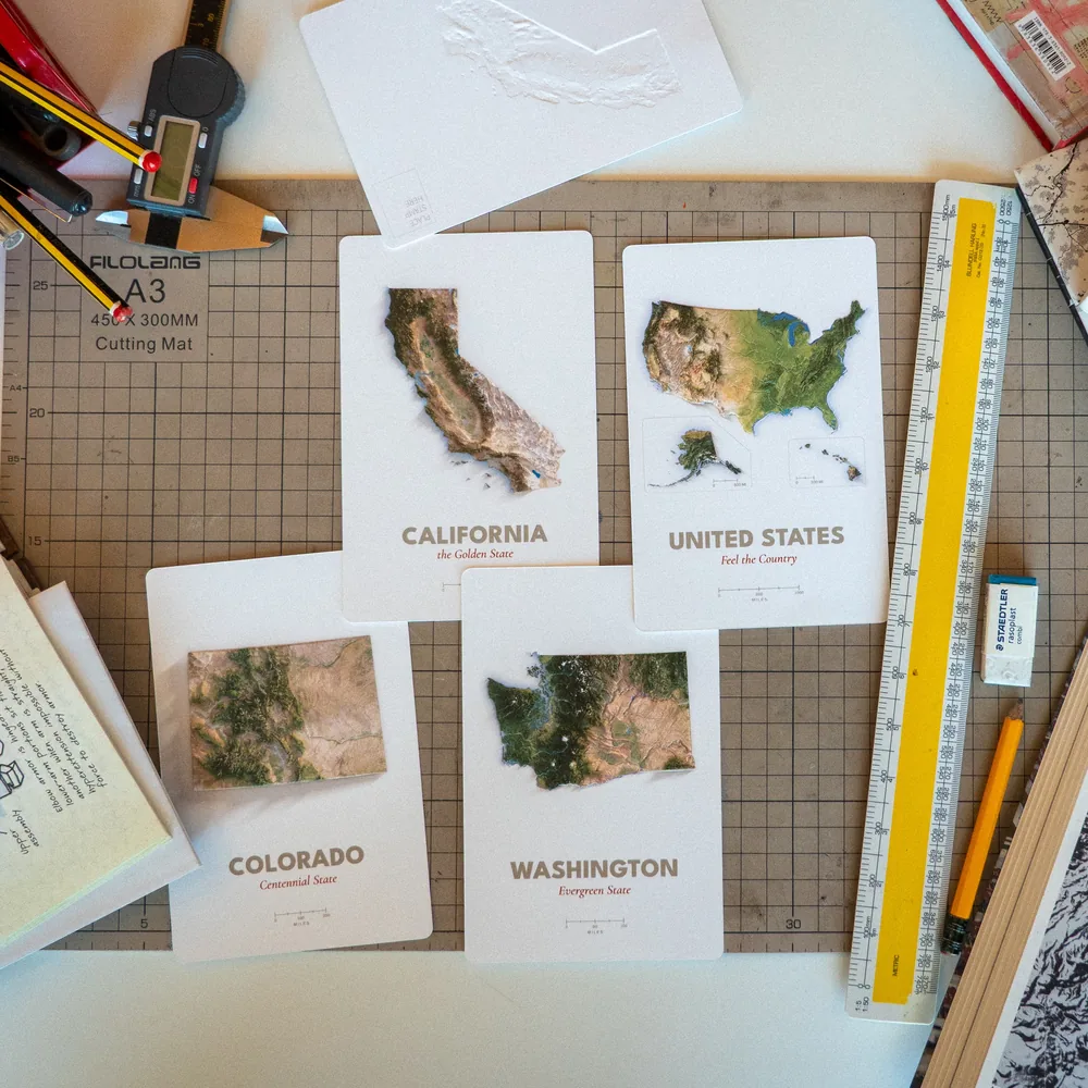 The California, United States, Colorado, and Washington proofs on a cutting mat with a ruler and pencils, one card turned over to show the relief from the back